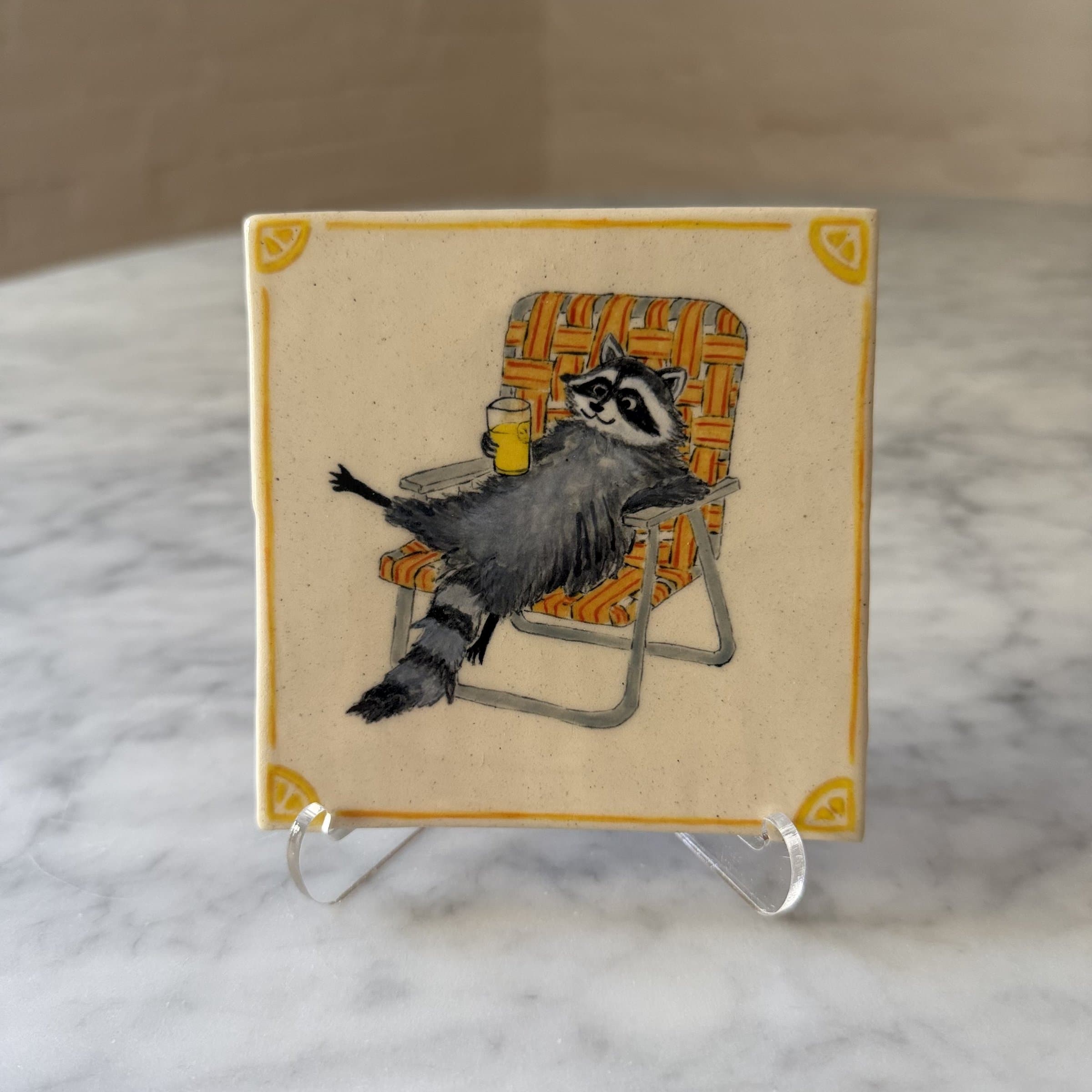 A hand-painted tile of a raccoon lounging in a striped lawn chair with a glass of lemonade.