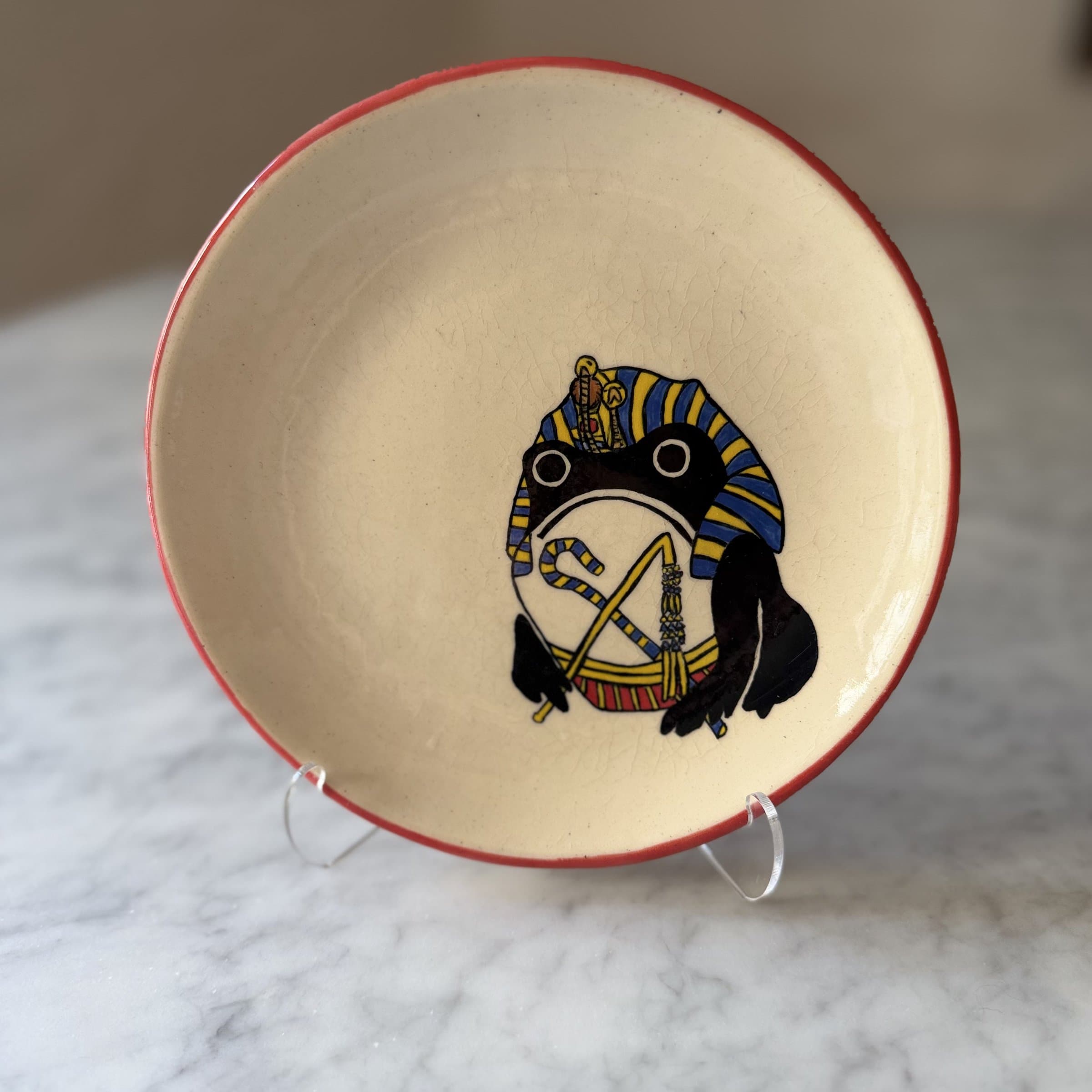 A plate painted with a frog wearing a striped pharaoh's nemes headdress and crook and flail.