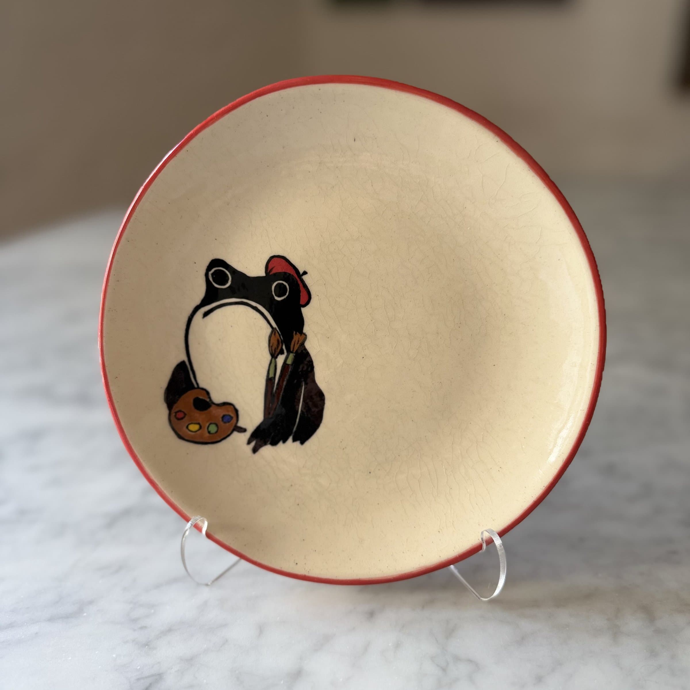 A plate painted with a frog wearing a red beret and holding a painter's palette and brushes.