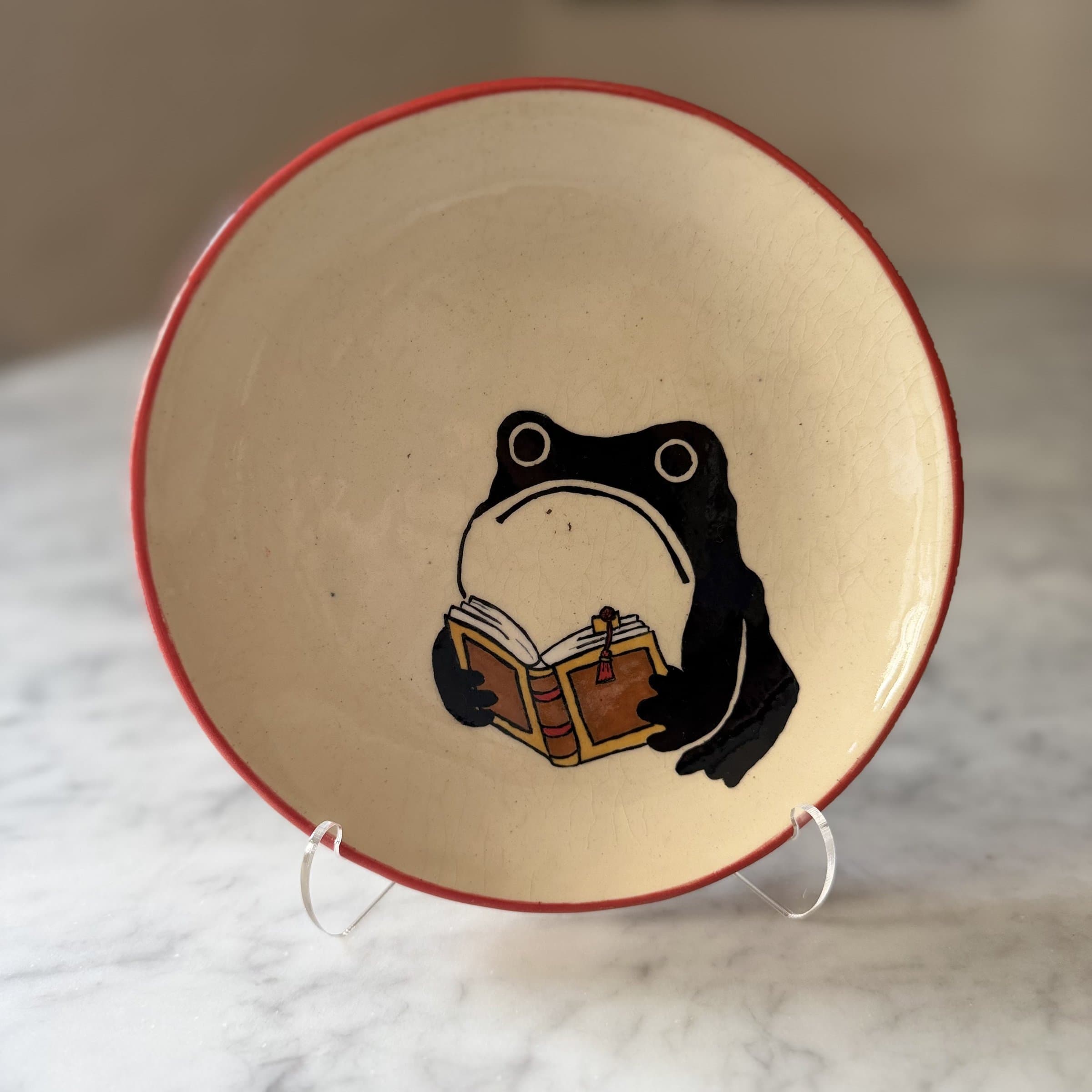 A plate painted with a frog holding open a hardbound book.