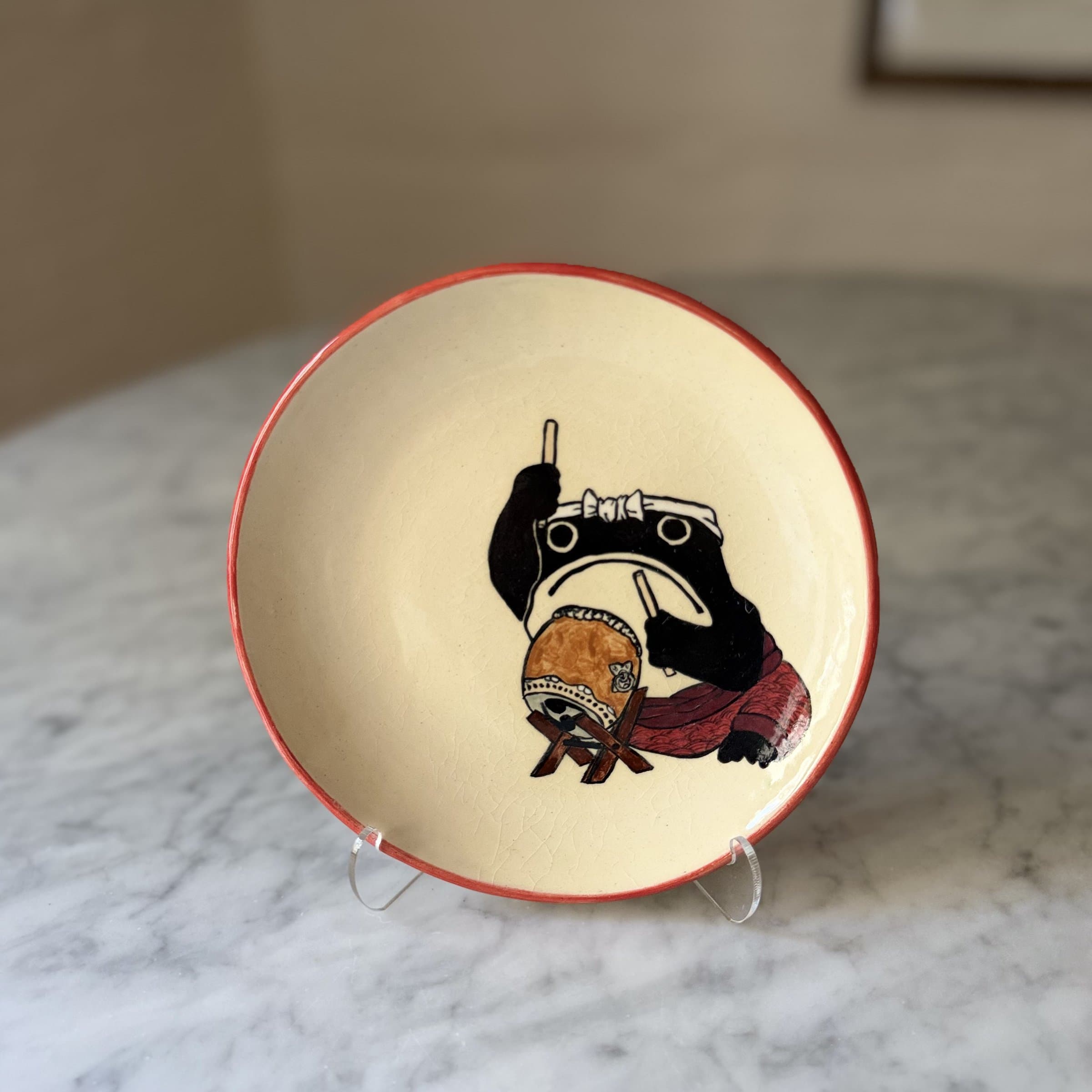 A plate painted with a frog playing a taiko drum with drumsticks raised.