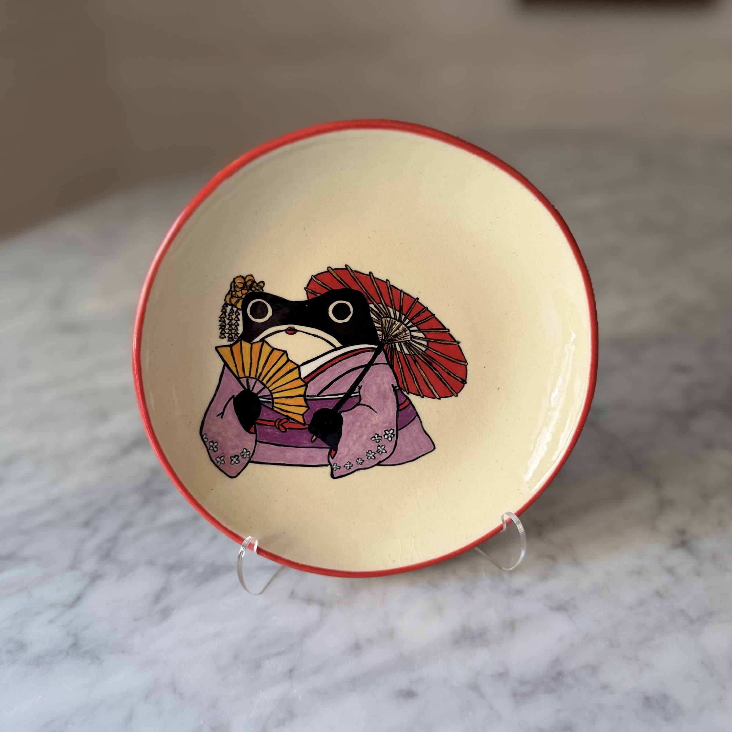 A plate painted with a frog in a purple kimono, holding a red parasol and yellow fan.