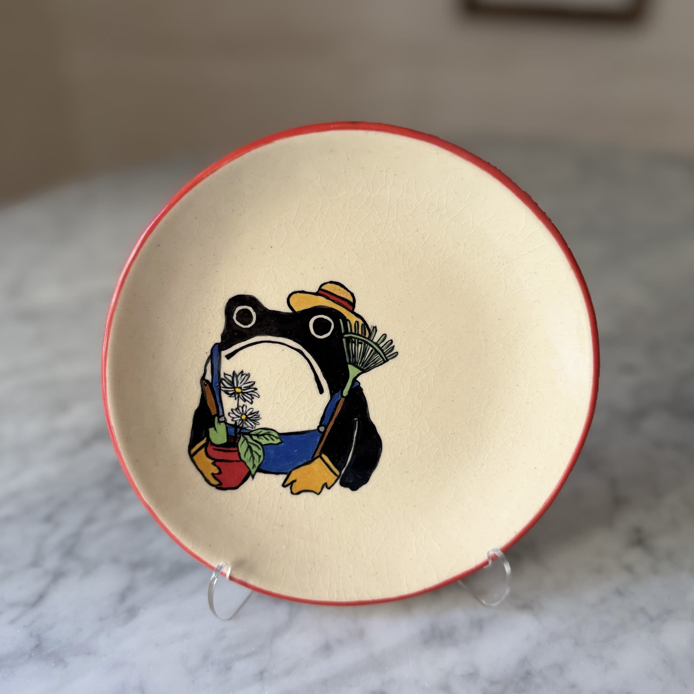 A plate painted with a frog in gardener's apron and straw hat, holding a rake and a potted flower.