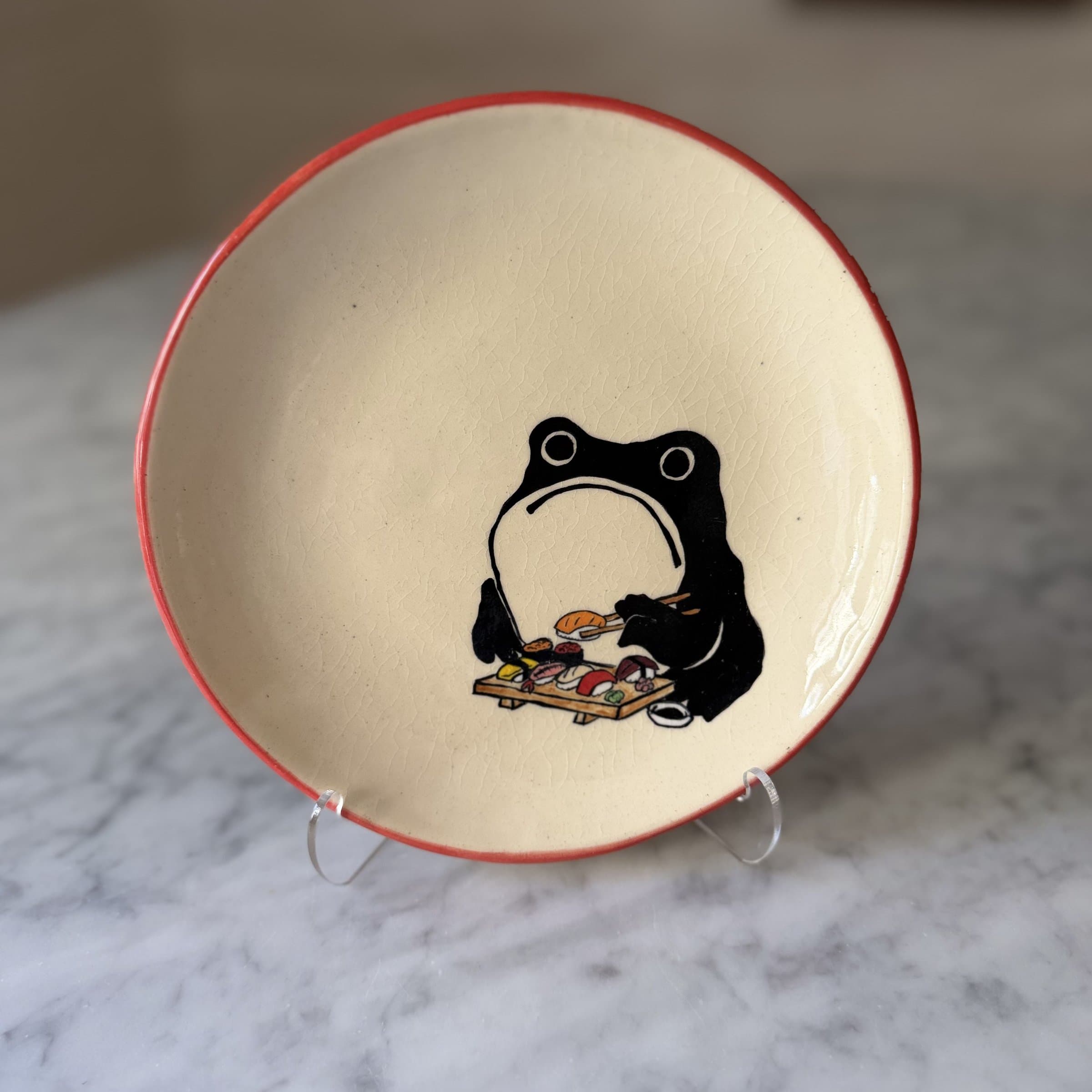 A plate painted with a frog holding chopsticks over a wooden board of sushi.