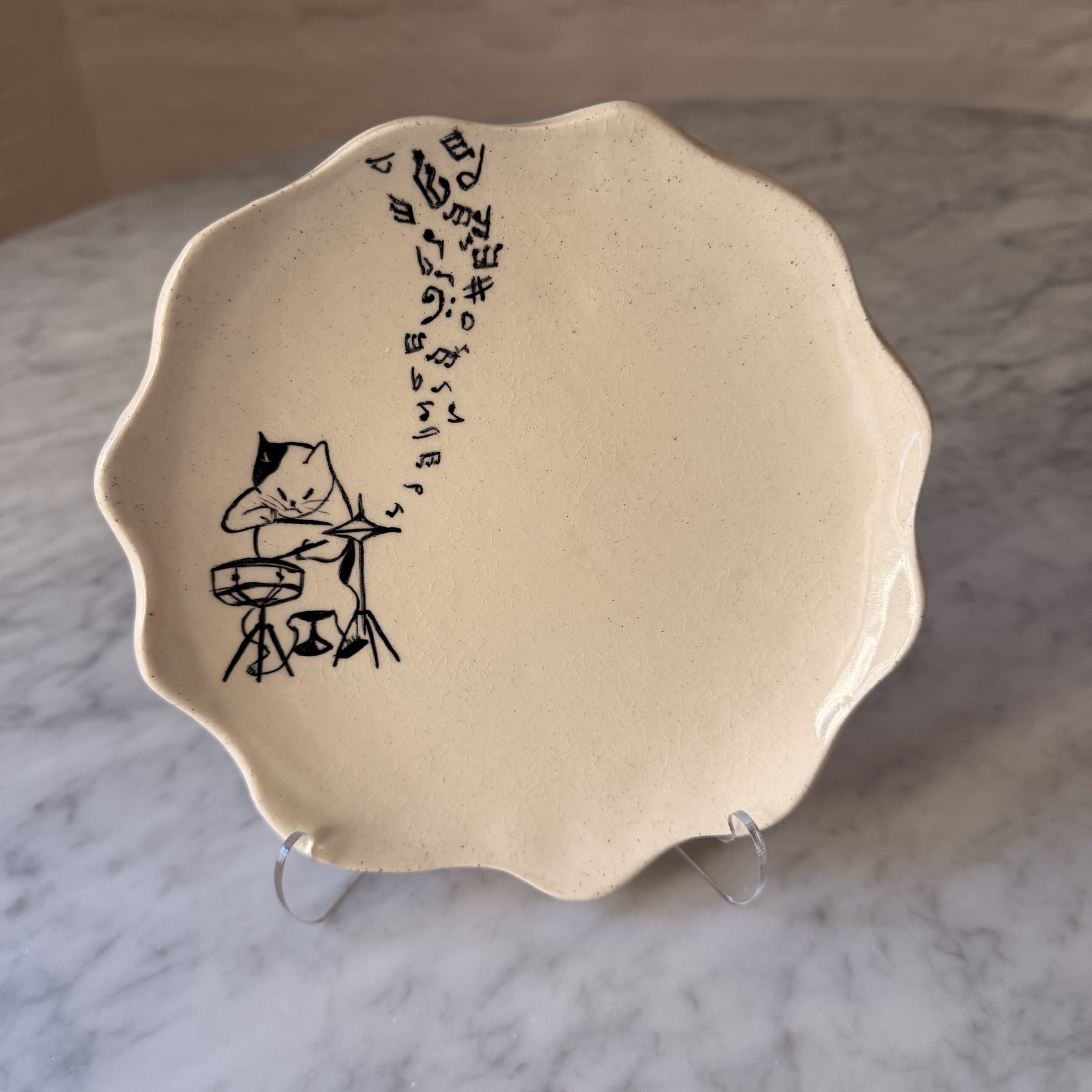 A scalloped plate painted with a cat playing a drum kit, music notes rising.