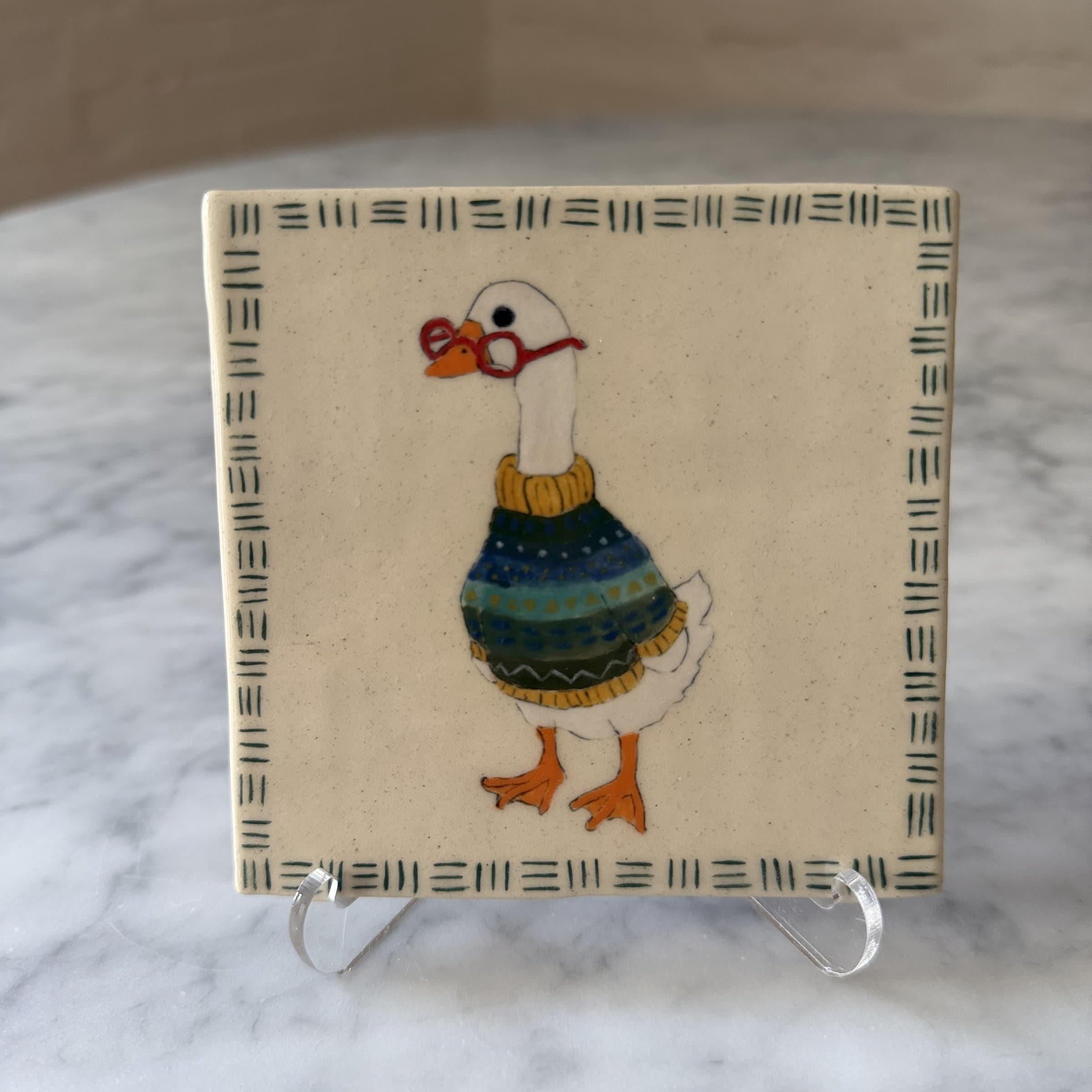 A hand-painted tile of a goose wearing reading glasses and a patterned sweater.