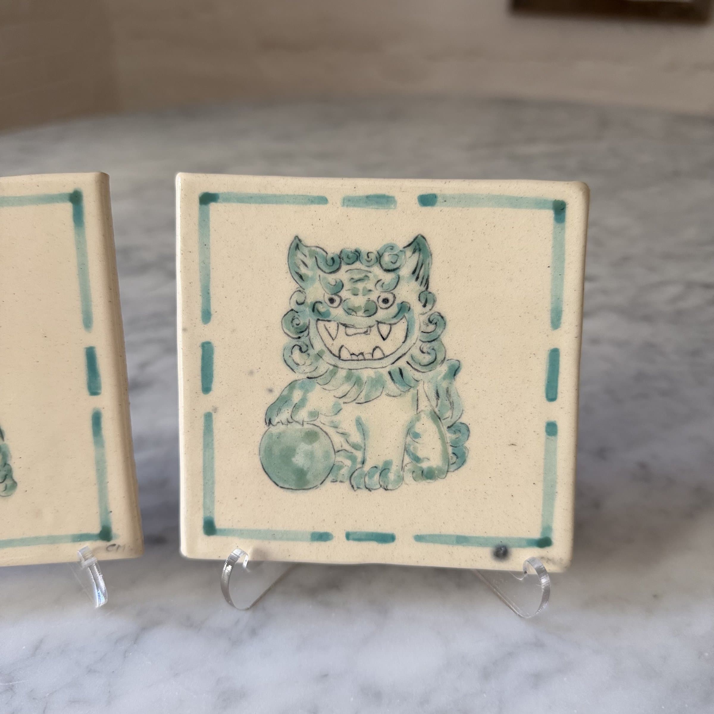A hand-painted tile of a fierce green foo dog with one paw on an embroidered ball.