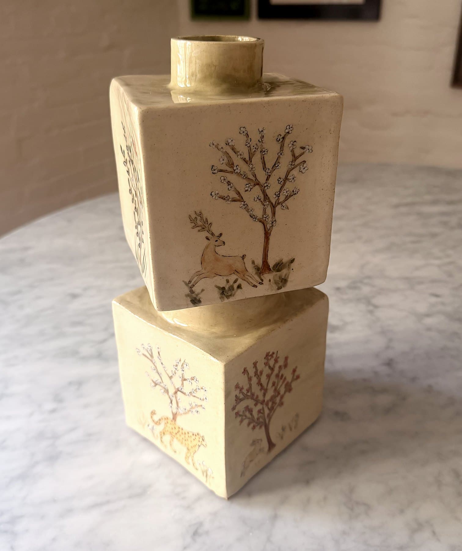 Two stacked, offset ceramic cubes painted with blossoming trees, a leaping deer and a leopard, turning slowly.