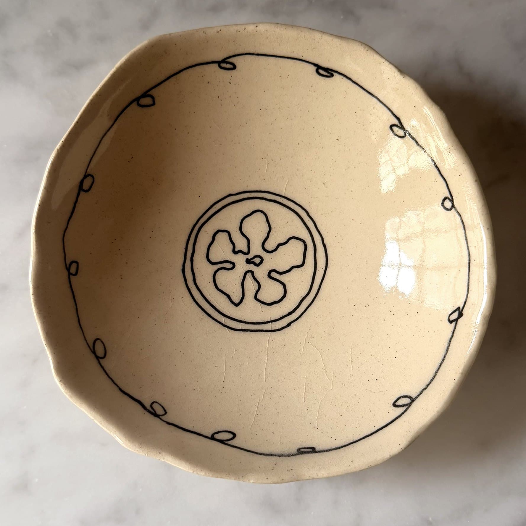 A hand-built cream bowl with a looped black border and a flower inside a double circle.