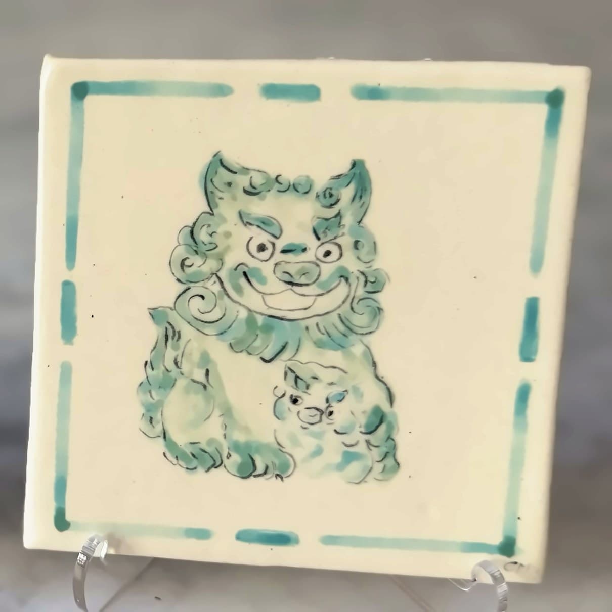 A tile painted in green with a smiling foo dog and its cub.