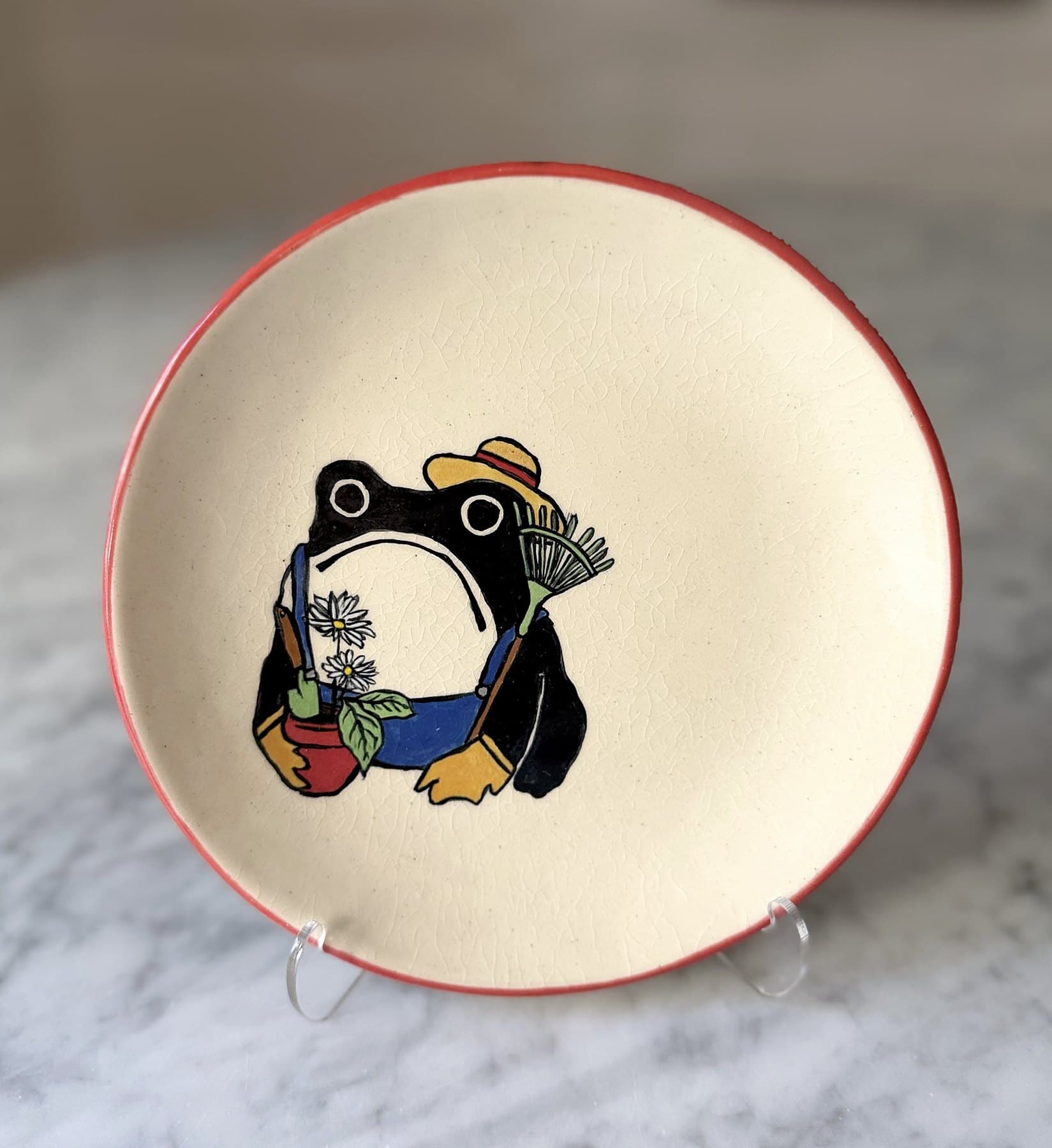 A red-rimmed plate painted with a frowning black frog in overalls and a straw hat, holding a potted daisy and a rake.