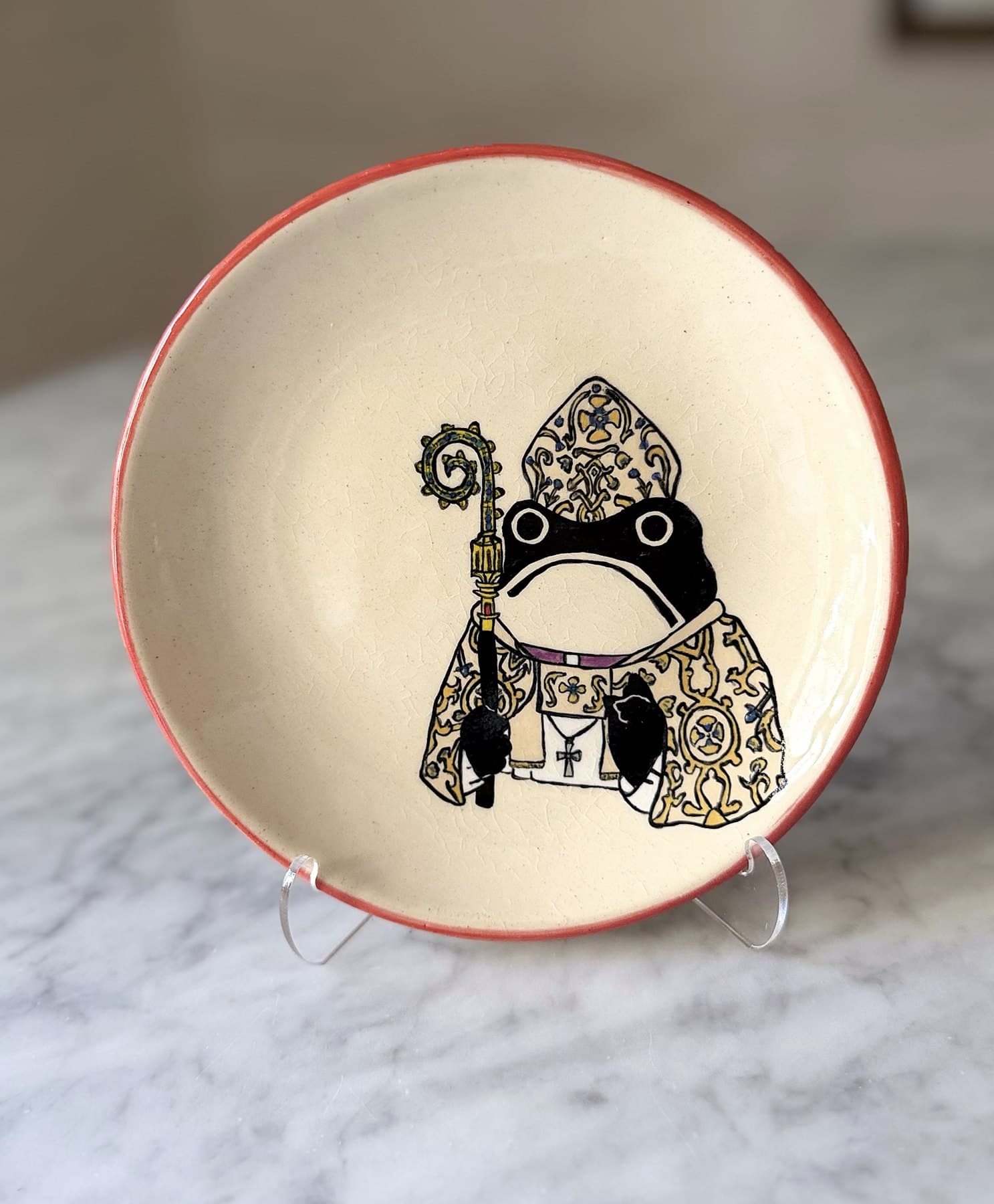 A red-rimmed plate painted with a frowning frog in a gold mitre and embroidered vestments, holding a crozier.