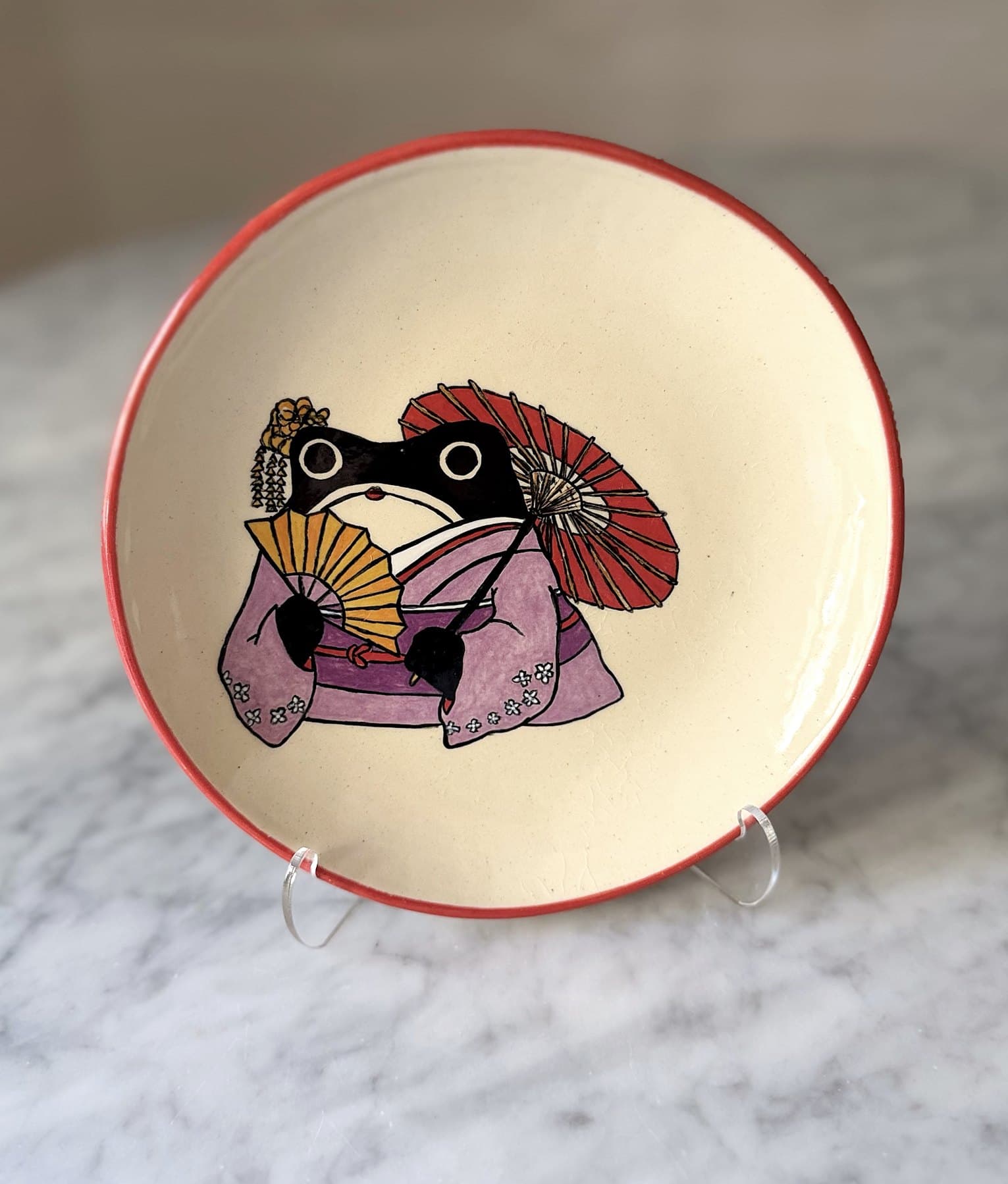 A red-rimmed plate painted with a frog in a lilac kimono holding a gold fan and a red paper parasol.