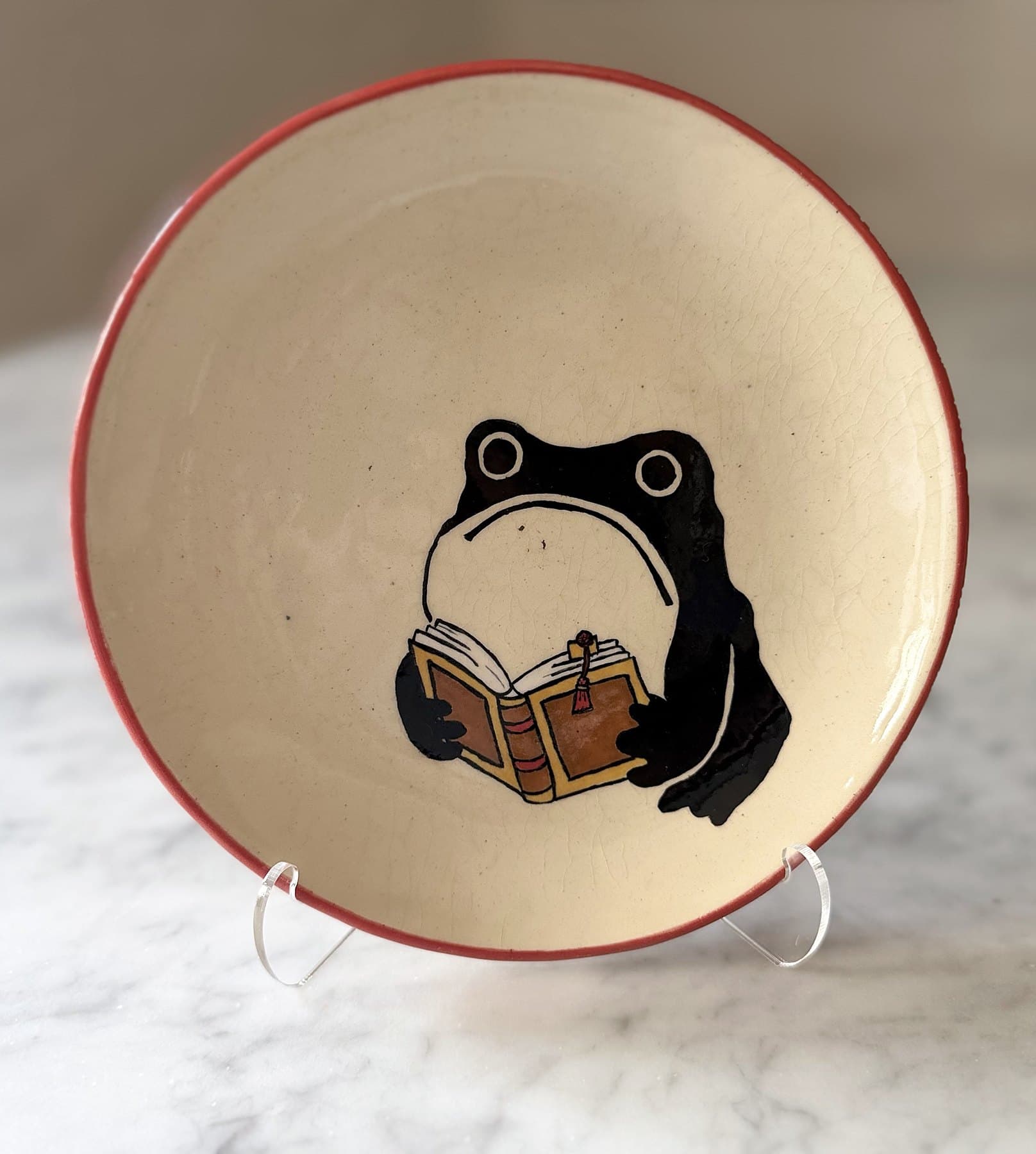 A red-rimmed plate painted with a frowning frog reading an open leather-bound book.