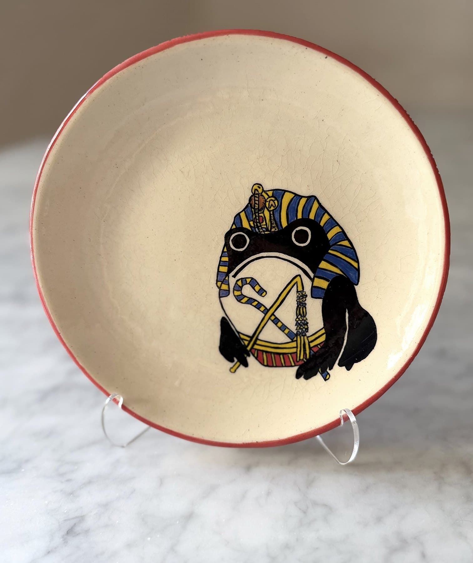 A red-rimmed plate painted with a frowning frog in a striped blue-and-gold pharaoh headdress, holding a crook and flail.