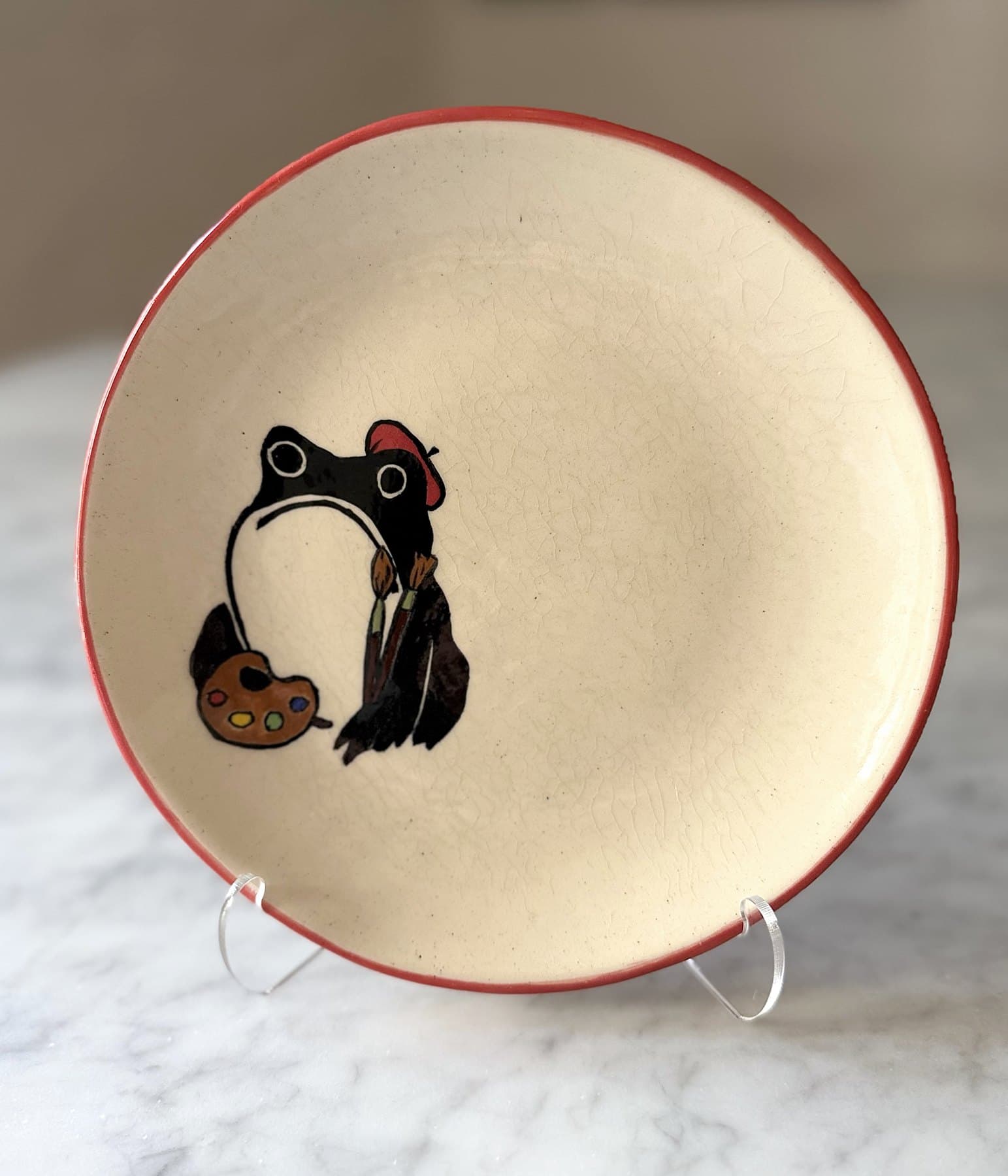 A red-rimmed plate painted with a frog in a red beret holding paintbrushes and a palette.